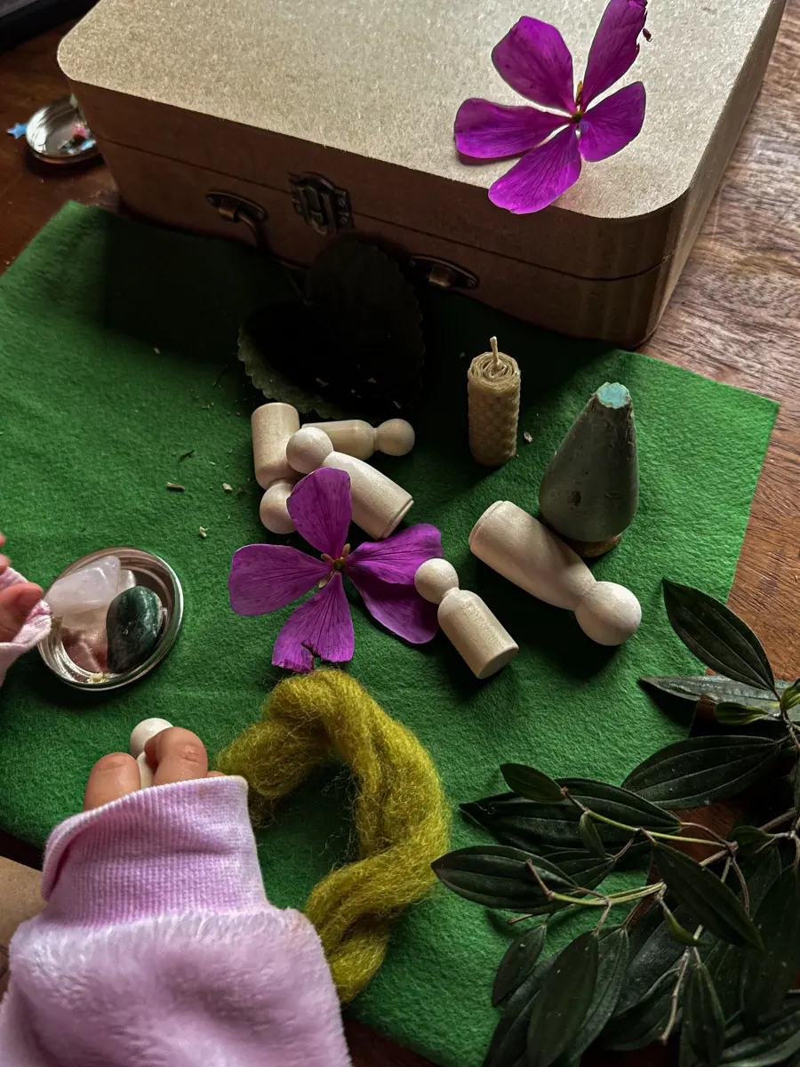 A maleta Pequena Terra fechada ao fundo e, à frente, mãos de criança brincando sobre o feltro com bonequinhos, vela e a árvore de madeira